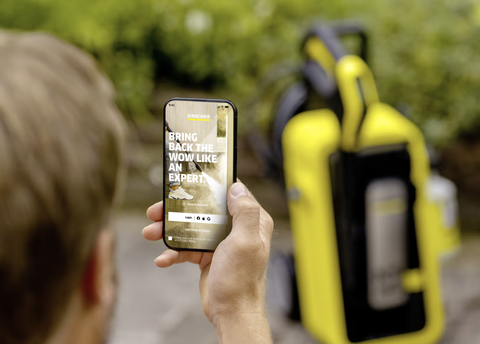 A user holding a smartphone showing the redesigned Kärcher Home & Garden app onboarding screen, with a yellow Kärcher pressure washer in the garden behind.